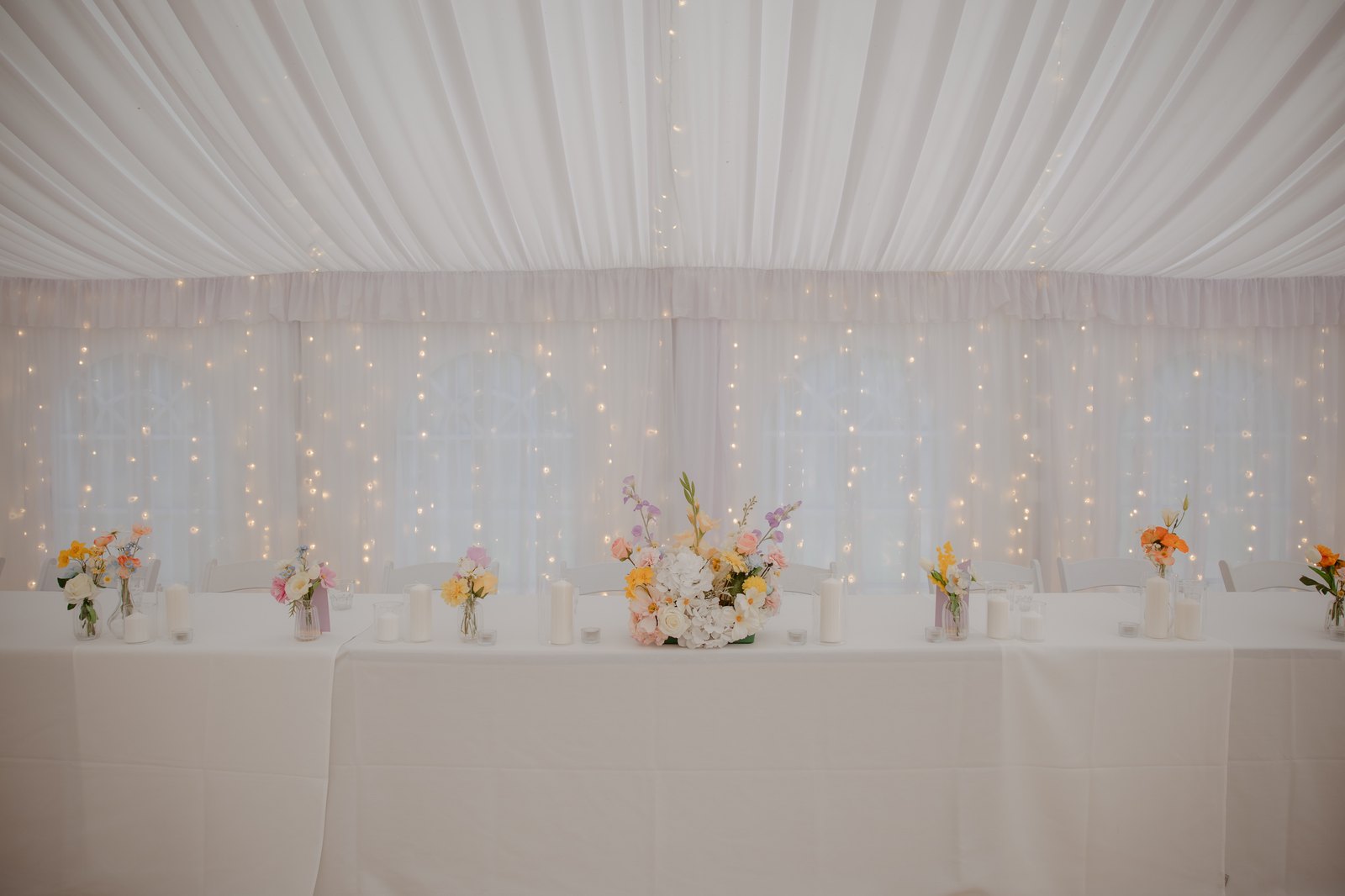 Long guest table under warm fairy lights, styled with bud vases and taper candles